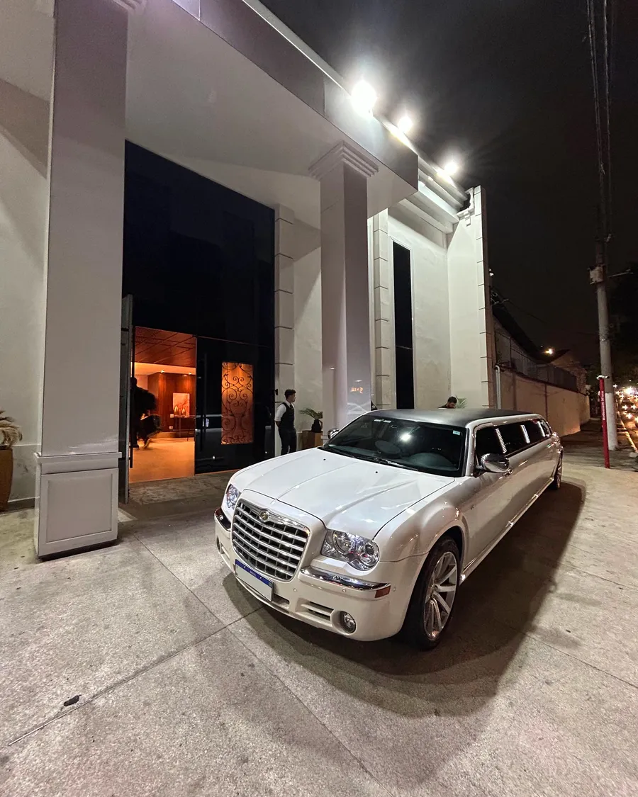 Limousine Chrysler 300C branca estacionada à noite na entrada de um edifício, com manobrista ao fundo
